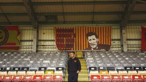 Image of Manpreet Singh at Firhill Stadium. He's standing in the middle of one of the stands with rows of empty grey, black, yellow and read seats to either side of him. There are three banners on the back wall. One reads "Johnnie Lambie's Red and Yellow". It has an image of Johnnie Lambie and over red and yellow stripes.