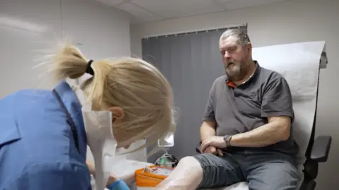 Malcolm Burns receives treatment to the wound on his leg. He is sitting in a patient's chair while a nurse tends to his wound. 