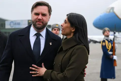 Getty Images the vances on a runway tarmac. ush has her arms around and is looking at jd who is looking towards the camera. there are soviet style army officers on the tarmac behind them