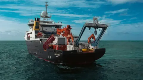 A boat with heavy machinery can be seen sailing on the sea. There are blue skies in the background. The ship has a black hull and there are orange cranes visible at the stern. 