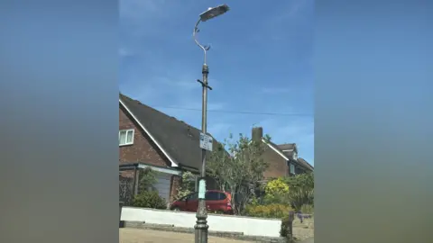 Katy Lewis/BBC A tall, older-style streetlamp with a curved arm stands on a quiet residential street, set against a clear blue sky. Behind it are brick houses with sloped roofs, a neatly kept garden, and a red car parked in a driveway. A small sign is attached to the lamp post, and the scene is brightly lit by midday sunlight.