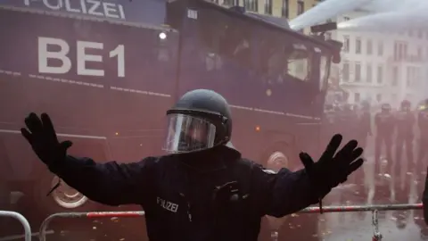 EPA A policeman raises his arms while riot police with a water cannon breaks up a demonstration against German coronavirus restrictions