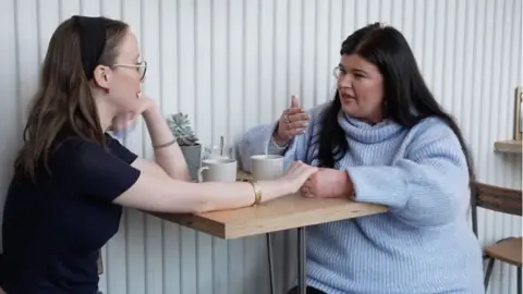 Kitty and Jessie sat at a table in a coffee shop. Kitty has long dark hair and is wearing a black headband. She has glasses and braces and is wearing a navy t-shirt. Jessie has long black hair and gold hoop earrings, and is wearing a light blue knitted roll neck jumper. 