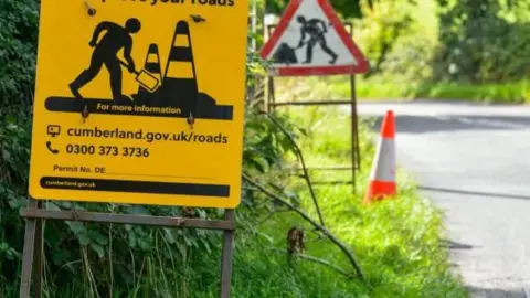A Cumberland Council yellow road sign which displays a work person digging at the side of the road. There is another highway sign in the distance and an orange traffic cone at the side of the grassy verge.