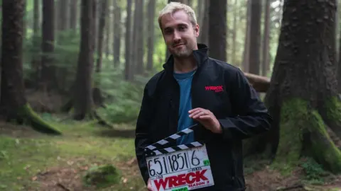 Ryan Brown Ryan Brown, pictured in woodland, with a clapperboard for Wreck season two. Ryan is a tall white man in his early 30s with blonde hair cut short and stubble. He wears a black waterproof jacket embroidered with 'Wreck Season 2' and a blue T-shirt.