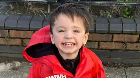 Merseyside Police Jude Gerrard - a young boy with brown hair, smiling and wearing a red waterproof coat with Marvel branding. 