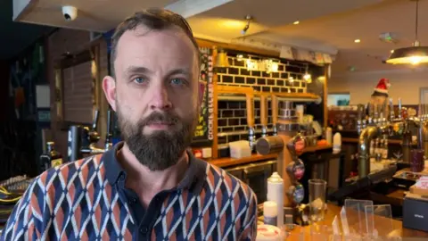 David stands in his now closed pub in Cardiff in front of the bar. He has short hair and blue eyes looking straight into the camera, and is wearing an orange and blue patterned polo shirt. Behind him is the bar where he used to serve customers now covered with boxes as they move equipment and decorations out.