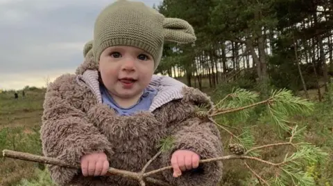 Steve Harris A baby dressed in woollen clothes and a pink woollen hat is holding a pine tree branch in a nature reserve. Other people could be seen in the background. It is overcast.