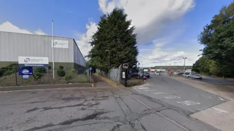 A road is prominent at the bottom of the photo. A building is on the left and two signs are visible in that part of the image including the word Wolverhampton on one of them. Trees are in the middle and on the right. 