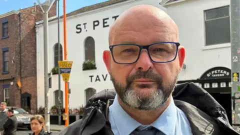 Frank Stevens, who aged around his late 40s and has a bald head, a grey and black beard and glasses, looks into the camera with a serious expression. He is wearing a black waterproof style coat and a blue shirt and tie. In the background is an orange lamp-post with a yellow sign, and pub with whitewashed brick walls. 