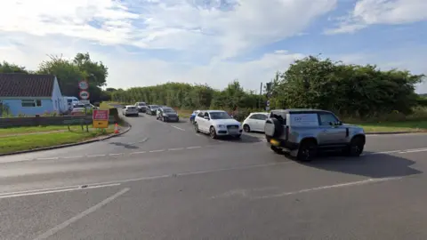 Google The Crostwick Lane junction, showing a queue of six cars looking to get on to the main road in the foreground. It is a sunny day and there is greenery on the right, and a blue coloured bungalow on the left of the junction.