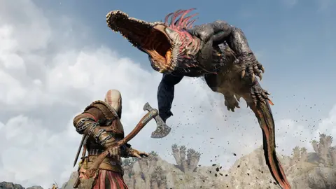 Sony A screenshot from God of War Ragnarök shows main character Kratos, viewed from behind, in a battle stance. He's readying his axe as a giant, crocodile-like creature leaps into the air, its jaws wide open, exposing rows of razor-sharp teeth as it prepares to attack our hero. Its long body is arched, and its long tail brushes against the ground, where a cloud of dust and debris has been kicked up as it left the ground moments before.