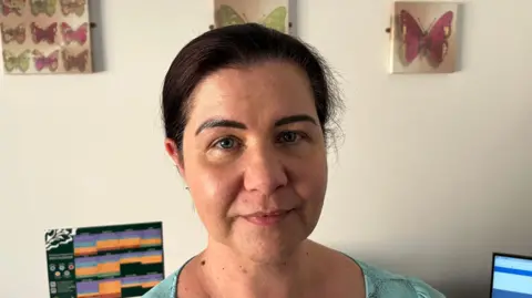 Meg Jones has black hair in a bun and is wearing a light green dress behind her are some canvass wall decorations of butterflies, a calendar can be seen too and part of a computer monitor.