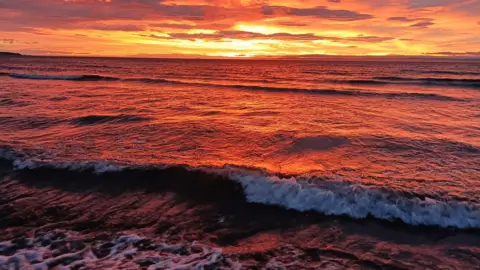 Shirley Faichney An orange and yellow sunrise appears through the clouds over some waves.