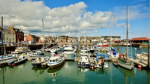 Graham Christie A view of a marina with lots of tall yachts and smaller boats on still water. Buildings can be seen along the far side of the pier.