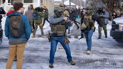 An officer with US Immigration and Customs Enforcement (right) and another federal officer holding a crowd control device (center) stand at a Minneapolis intersection where protesters had gathered after the death of Renee Good. Bystanders and journalists also stand in the intersection.