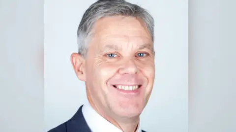 Cambridgeshire Fire and Rescue Matthew Warren wearing a dark suit, white shirt and tie looking towards the camera. He has grey hair and bright blue eyes. He is standing against a plain light blue background.