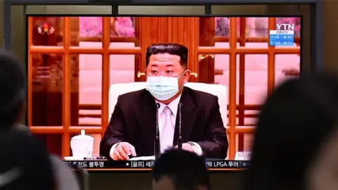 Getty Images People sit near a screen showing a news broadcast at a train station in Seoul on May 12, 2022, of North Koreas leader Kim Jong Un appearing in a face mask on television for the first time to order nationwide lockdowns after the North confirmed its first-ever Covid-19 cases.