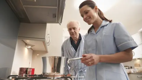 Care worker helps man cook in a kitchen, with pan on a hob