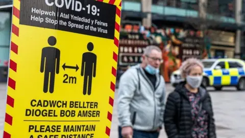  Couple in face masks walk along high street next to a Covid-19 warning sign advising people to maintain a distance from each other with a police car in the background