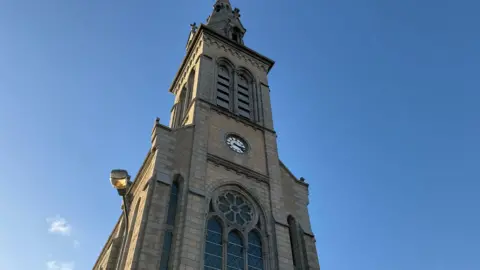 The outside of a church tower on a clear sunny day. The camera is looking up at the church clock tower. There is clear blue skies with only a few clouds in the sky. The church has a clock and some large windows. 