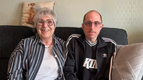 a picture of wayne who's wearing a black tracksuit top, shirt, glasses and short hair.
His mum karen is on the left wearing a striped shirt over a white blouse, she has short-ish white hair and hexagonal glasses - she's smiling a broad smile.
