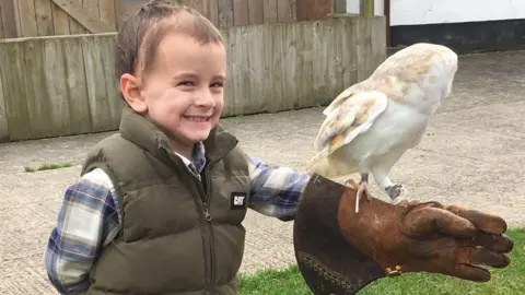 Claire Carrick A young boy with short dark hair, a green waistcoat and a long-sleeved shirt, smiles while a white owl is perched on a large brown glove he is wearing on his left hand. 