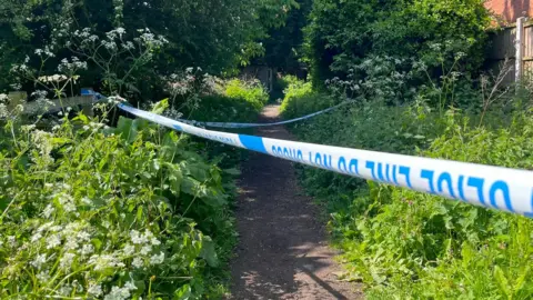 Police tape blocks part of a muddy footpath which has weeds and bushes either side. There is a fence to the right hand side of the image with a building behind that.