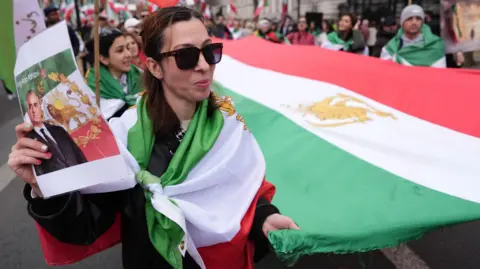 PA Media A woman smiles as she and others hold the sides of a large version of the Iranian flag with a Persian symbol in the centre. She also holds a printout of former Shah (king) Reza Pahlavi.