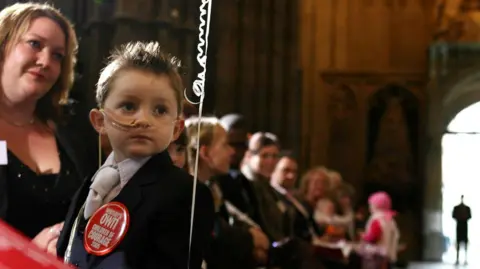 PA Media A mother and child in a church, the child is a young boy who has a tube in his nose and a red badge which reads 'Women's Own Children of Courage'
