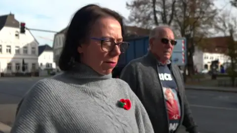 Natasha Walker walks in a street beside her father Richard Lee. She had brown, shoulder-length hair and glasses and wears a poppy badge on a light-grey sweater. He has grey hair and wears sunglasses and a sweatshirt with a picture of Katrice in a red dress.