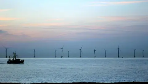 A fishing boat in the sea in silhouette with offshore wind turbines in the background. The picture has been taken on a summer evening. The sky is blue and the clouds have a pink tinge.