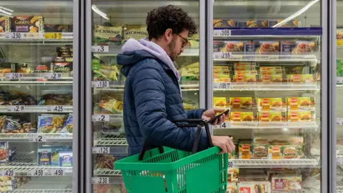 Getty Images Man in frozen food section of supermarket looking at prices