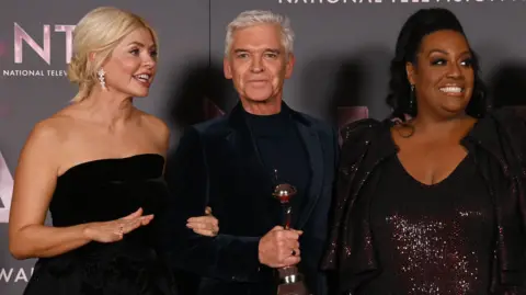 Getty Holly Willoughby, wearing a black strapless evening dress, stands holding an award with Phillip Scofield, center, and Alison Hammond, right, wearing a maroon shiny dress and jacket. 