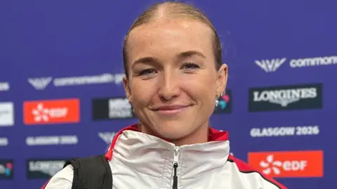 Lucy Woodward is wearing a white and red Team Jersey tracksuit jacket. She stands in front of a purple Commonwealth Games Glasgow 2026 media backdrop covered with event and sponsor logos. 