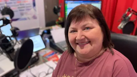 A woman with chin length brown hair is smiling at the camera. She is wearing a plum coloured t-shirt and is sitting in front of a radio studio