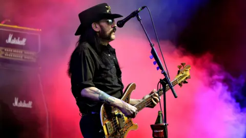 Lemmy while performing on stage with Motorhead. He has long black air and is wearing a black hat, black shirt with the sleeves rolled up and black trousers. He is standing facing to the right next to a microphone and is holding a bass guitar. A Marshall guitar amplifier and cabinet is visible on the left as pink/purple smoke rises behind him.