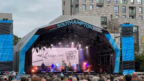 Dan Kerins A stage in an urban setting, with the Kaiser Chiefs onstage in front of a large video screen with tower blocks in the background and a crowd of people in the foreground