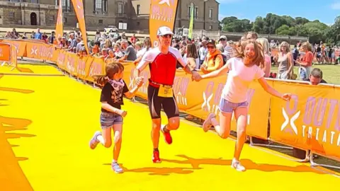 Alison Watson Neal Watson crossing the finish line of the Outlaw Half in Holkham, Norfolk, with his two daughters