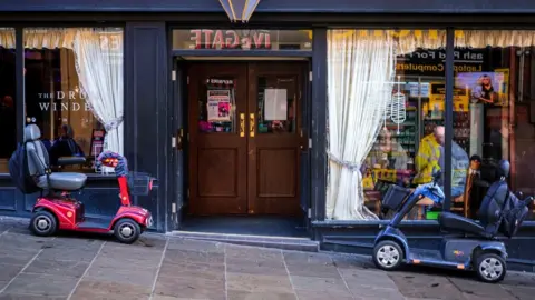 Marcus Rattray/Historic England 'Pub Lunch' - Two mobility scooters parked outside the Drum Winder public house. Drum Winder Public House, 12-12A Ivegate, Bradford.