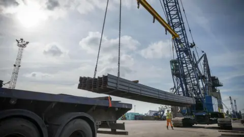 Large steel rods are lifted with a crane. It is a sunny day with blue skies. 