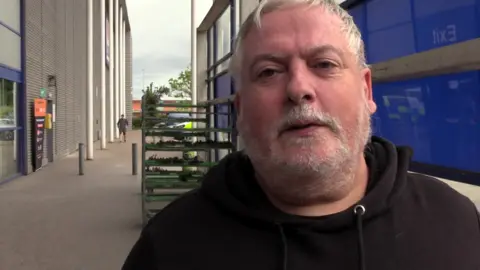 Simon Price is wearing a black hooded top outside what looks like a garden centre.