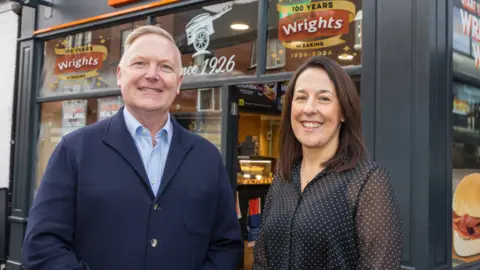Wrights Peter and Helen stand outside a Wrights shop which is black with orange livery and adverts. Peter is wearing a navy shirt and cardigan and has short grey hair and Helen has a black top on with shoulder length hair.