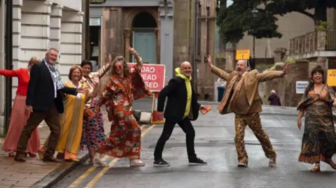 Jonathan Macfarlane  A group of people all wearing bright and unusual clothes walking across the road.