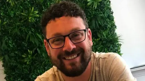 Jamie Wrighton A man with short brown curly hair, a brown beard and glasses smiles. He wears a grey t-shirt. Behind him, a fake plant wall can be seen.