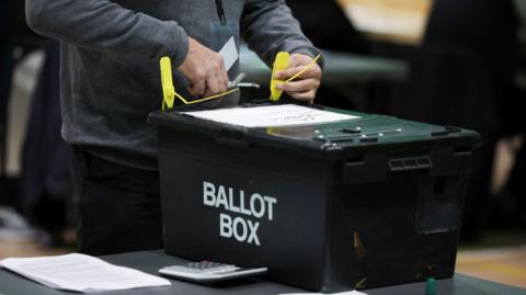 A person in dark clothes securing a ballot box with yellow plastic locks, with a set of papers and calculator to the right of the box.
