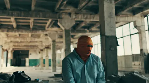 Ben Steele/BBC A former Russian army officer, who we have not named, sits in a warehouse in an orange balaclava.