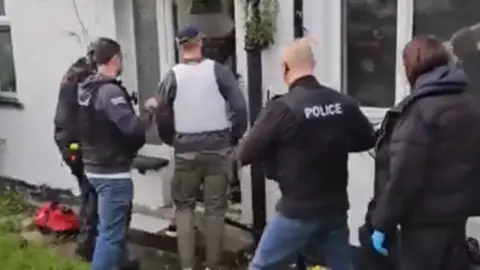 The backs of police officers in black stab vests going through a white PVC front door of a home. 