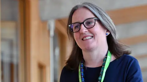 Getty Images Maree Todd walking in the Scottish Parliament. She is wearing glasses and is wearing a blue top, a blue and green necklace, and a green lanyard