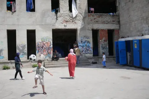 BBC A young boy plays with a ball in front of the building, which has no doors or windows - a few portable toilets stand against a nearby wall